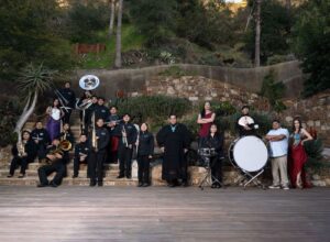 A group of performers from Al Sur de la Frontera stand outdoors in front of a small hill. Some carry instruments.