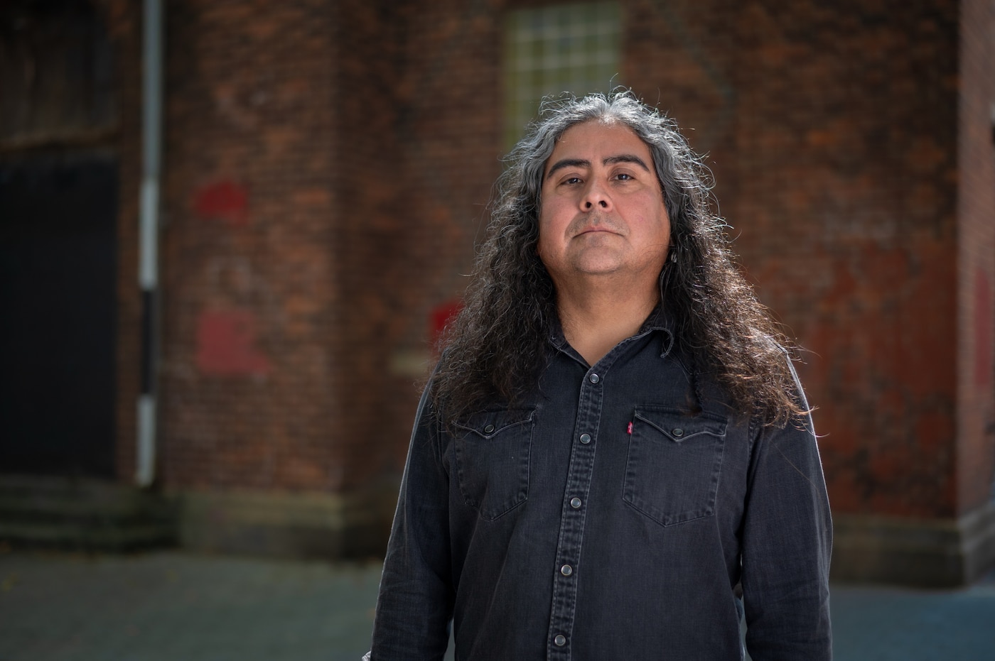 An image of artist and composer Raven Chacon standing in front of a building looking straight at the camera.
