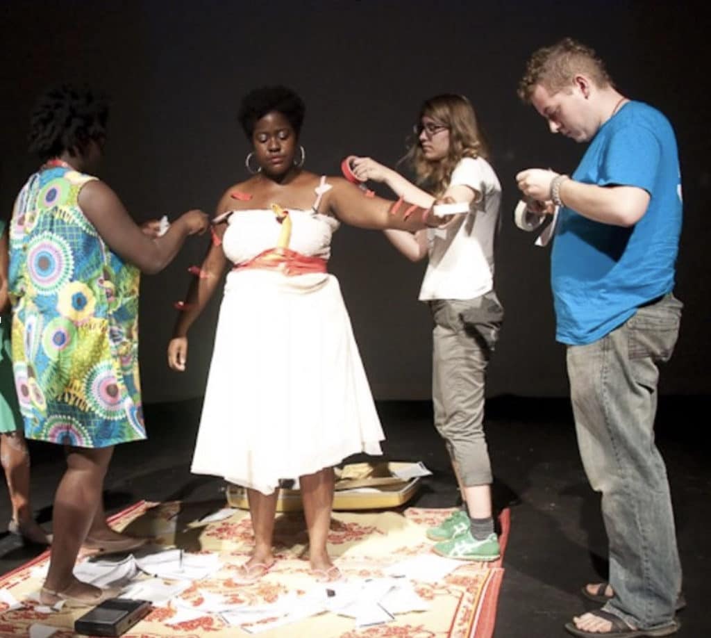 Gabrielle Civil (second from left) stands in white dress with red sash as three other people attach pieces of red and white tape to her arms