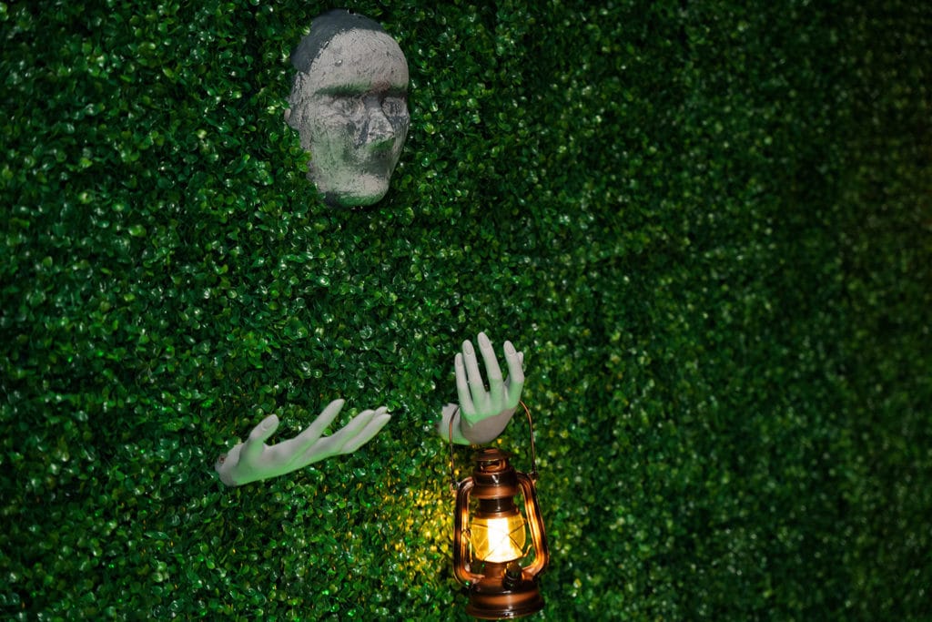 Stone face and hands holding a lantern emerge from foliage wall.