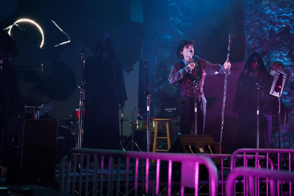 Student in vest and hat performs on stage, with cloaked figures standing behind them.