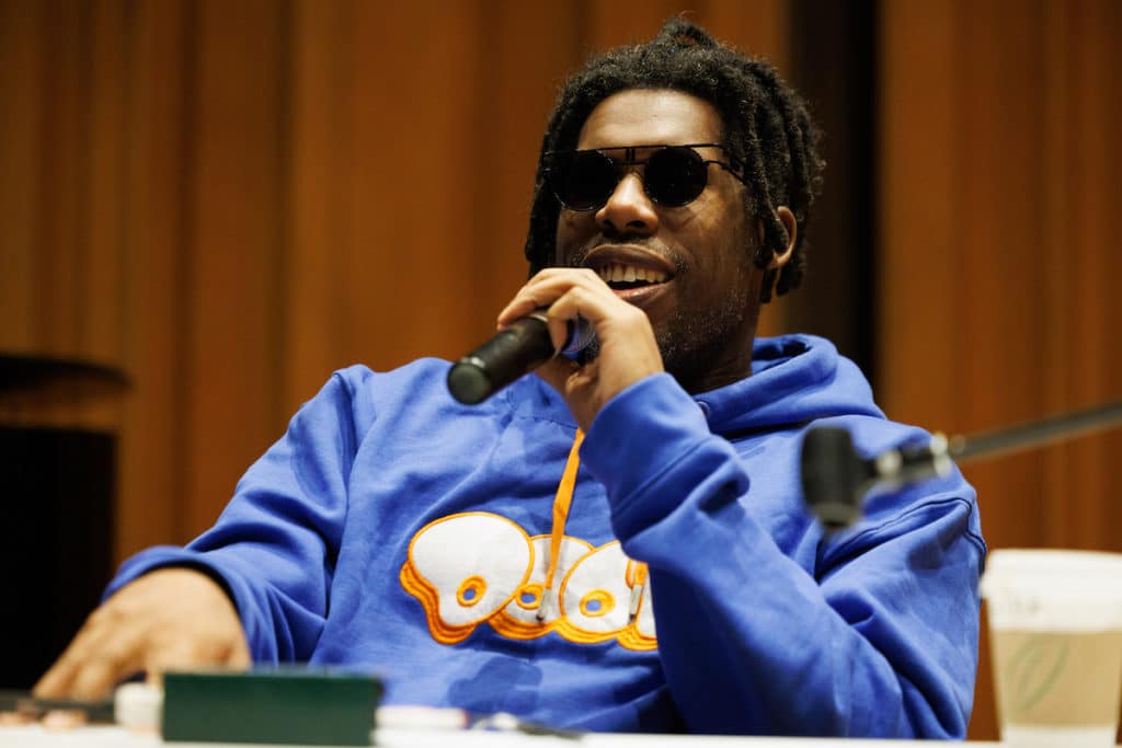 Flying Lotus, wearing sunglasses and a blue hoodie, speaks into a mic at a visiting artist talk in the ROD at CalArts.