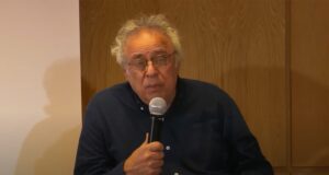 Norman Klein wearing glasses and navy blue shirt holding a microphone