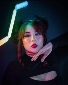 Woman in black top and colorful makeup poses with hand under chin, standing under multicolor lights