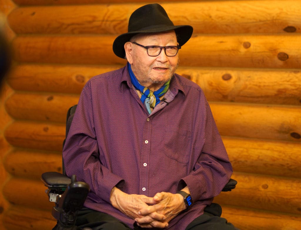 Writer N. Scott Momaday in wheelchair, sitting against a wall akin to a log cabin.