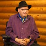 Writer N. Scott Momaday in wheelchair, sitting against a wall akin to a log cabin.
