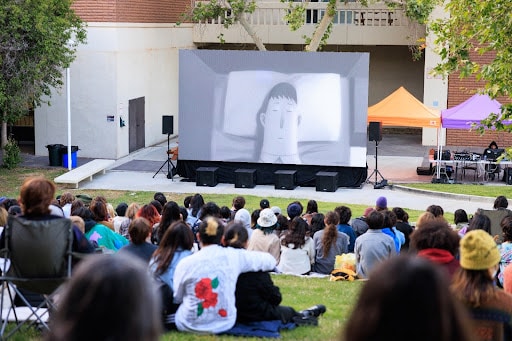 A group sits on the lawn outside, watching an animated film on a large screen.