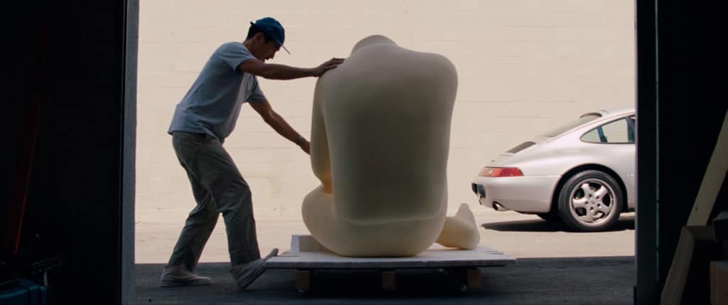 Artist supporting back of a sculpture of a kneeling figure with car in the background