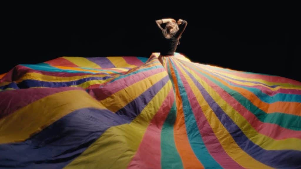 Julia Holter looks upward with arms raised, wearing massive colorful striped tarp as skirt