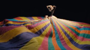 Julia Holter looks upward with arms raised, wearing massive colorful striped tarp as skirt