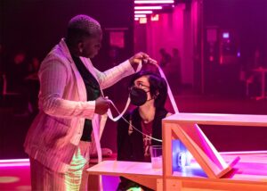 Autumn Knight, wearing striped white suit, holds long tape around the head of a masked participant