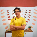 Daniel Corral, wearing yellow T-shirt, stands with arms folded in front of abstract mural and table