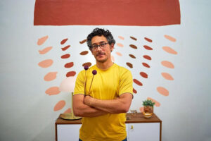 Daniel Corral, wearing yellow T-shirt, stands with arms folded in front of abstract mural and table