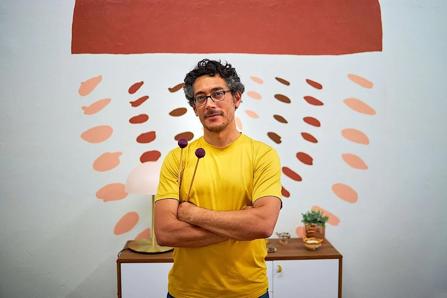 Daniel Corral, wearing yellow T-shirt, stands with arms folded in front of abstract mural and table