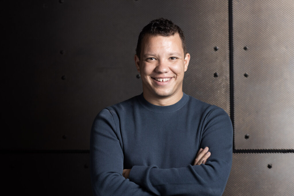 A headshot of Ryan Bancroft in a blue shirt against a beige stone wall.