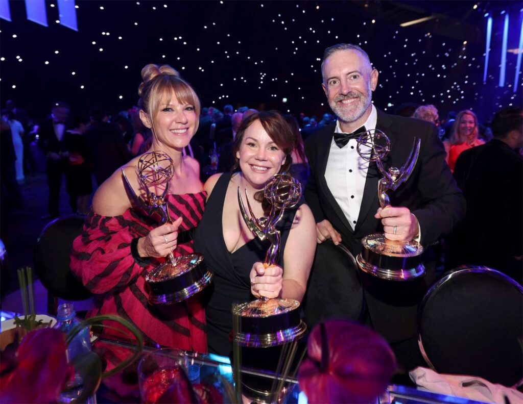 Three individuals in a theater smile at the camera, holding their Emmy Awards.