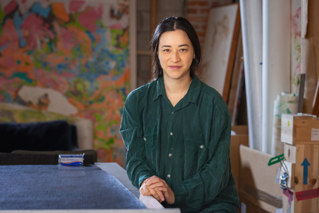 A portrait of artist Gala Porras-Kim in a green shirt in a studio or workspace.