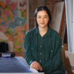 A portrait of artist Gala Porras-Kim in a green shirt in a studio or workspace.