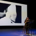 A woman standing in front of a podium on a large stage with a large screen displayed behind her showing a black and white image