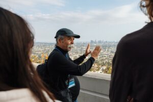 Harry Gamboa Jr. wearing a ball cap frames the skyline of LA with his hands while talking to a class of students.