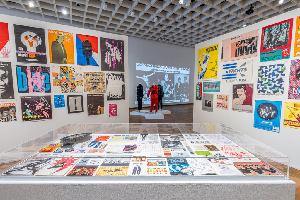 Glass case of punk memorabilia surrounded by walls plastered with punk posters, with clothing on mannequins in the distance