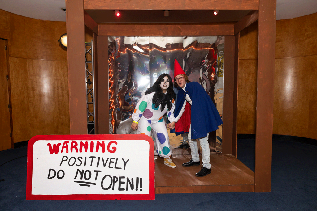 Two students in costume hold hands in a life-size box with large sticker reading 'Warning positively do not open'