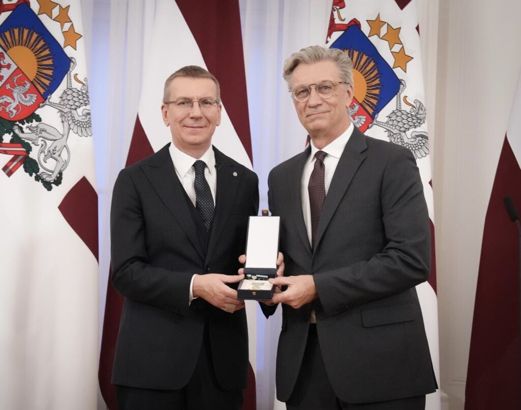 Two men in suits pose with a medal in front of official Latvian flags.