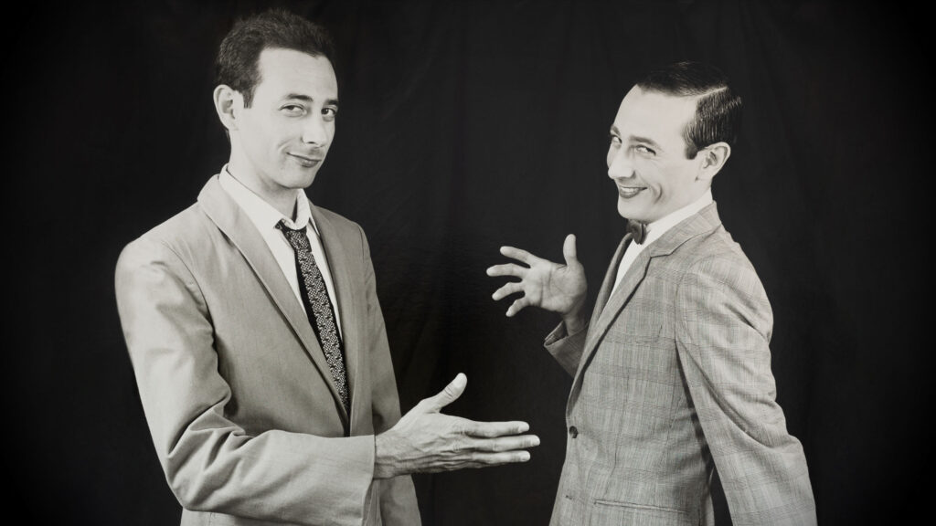 Paul Reubens and his character Pee-Wee Herman gesturing at a handshake