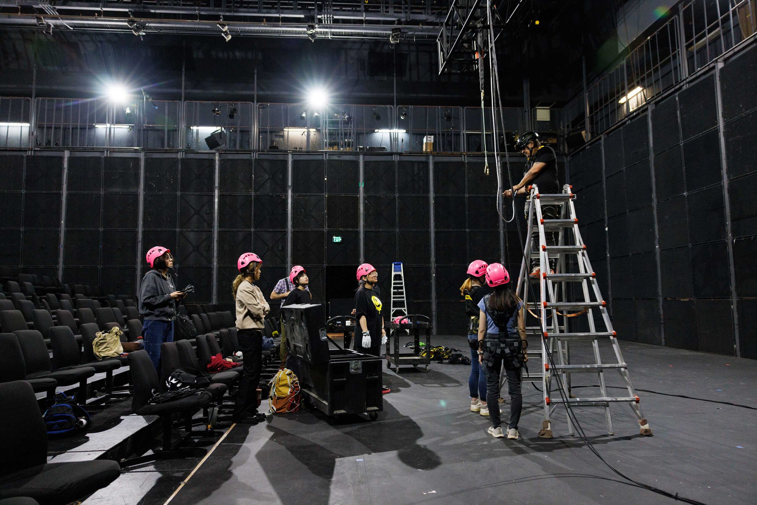 A group of student stand wearing hard hats in a large theater, as an instructor stands on a ladder with ropes and a harness