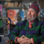 Peter Shire, smiling, sits in colorful studio with hands clasped