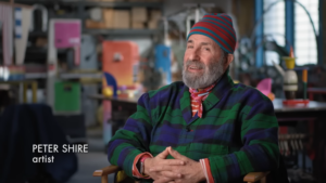 Peter Shire, smiling, sits in colorful studio with hands clasped