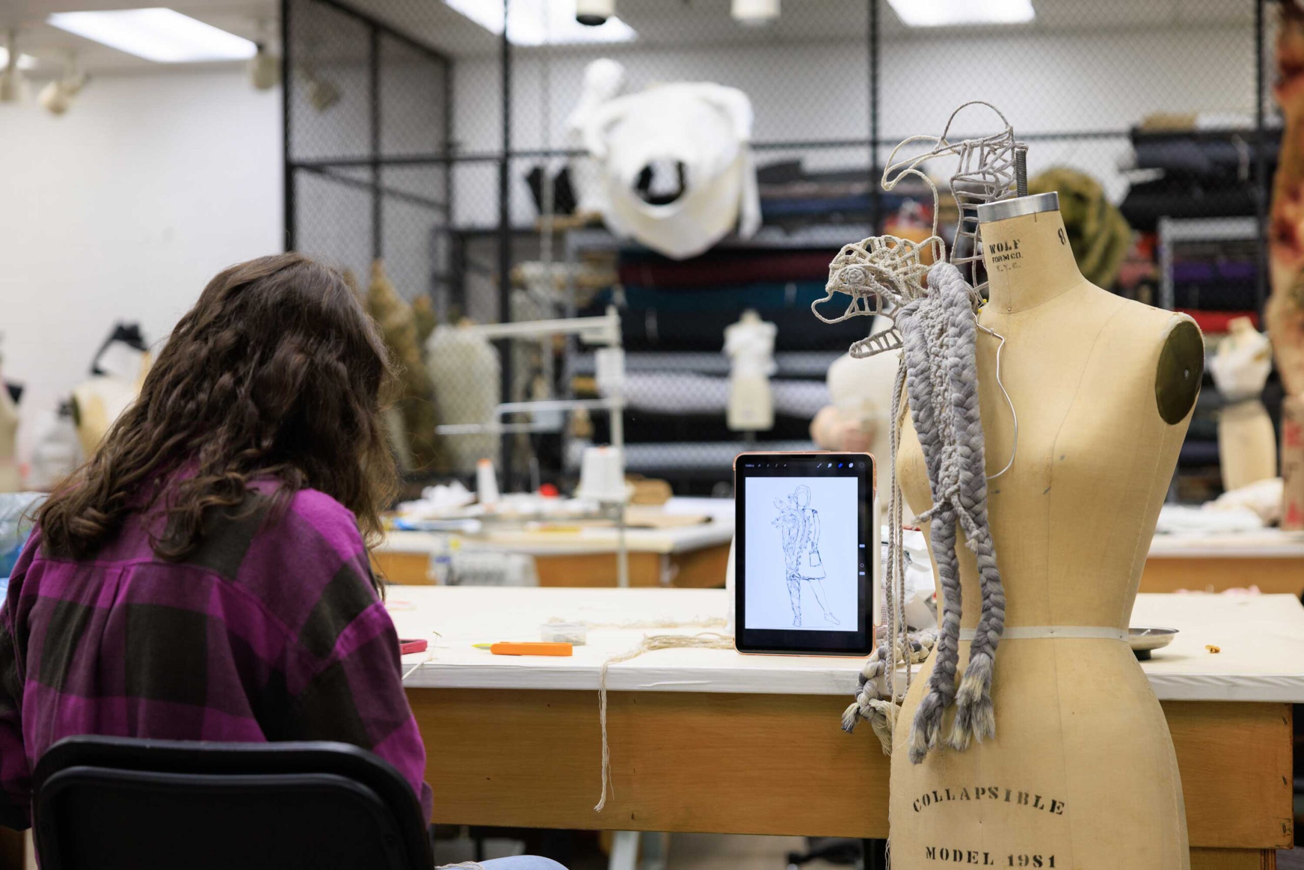 A student sits intently working next to their costume in process while an iPad shows a sketch of the final vision