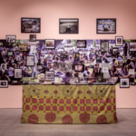 Installation view of table with decorate gold and red cloth in front of large photo collage