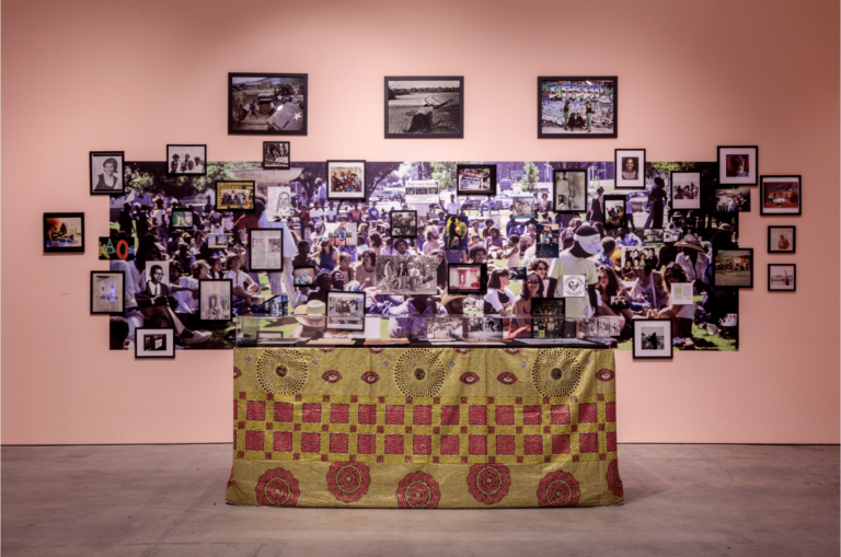 Installation view of table with decorate gold and red cloth in front of large photo collage