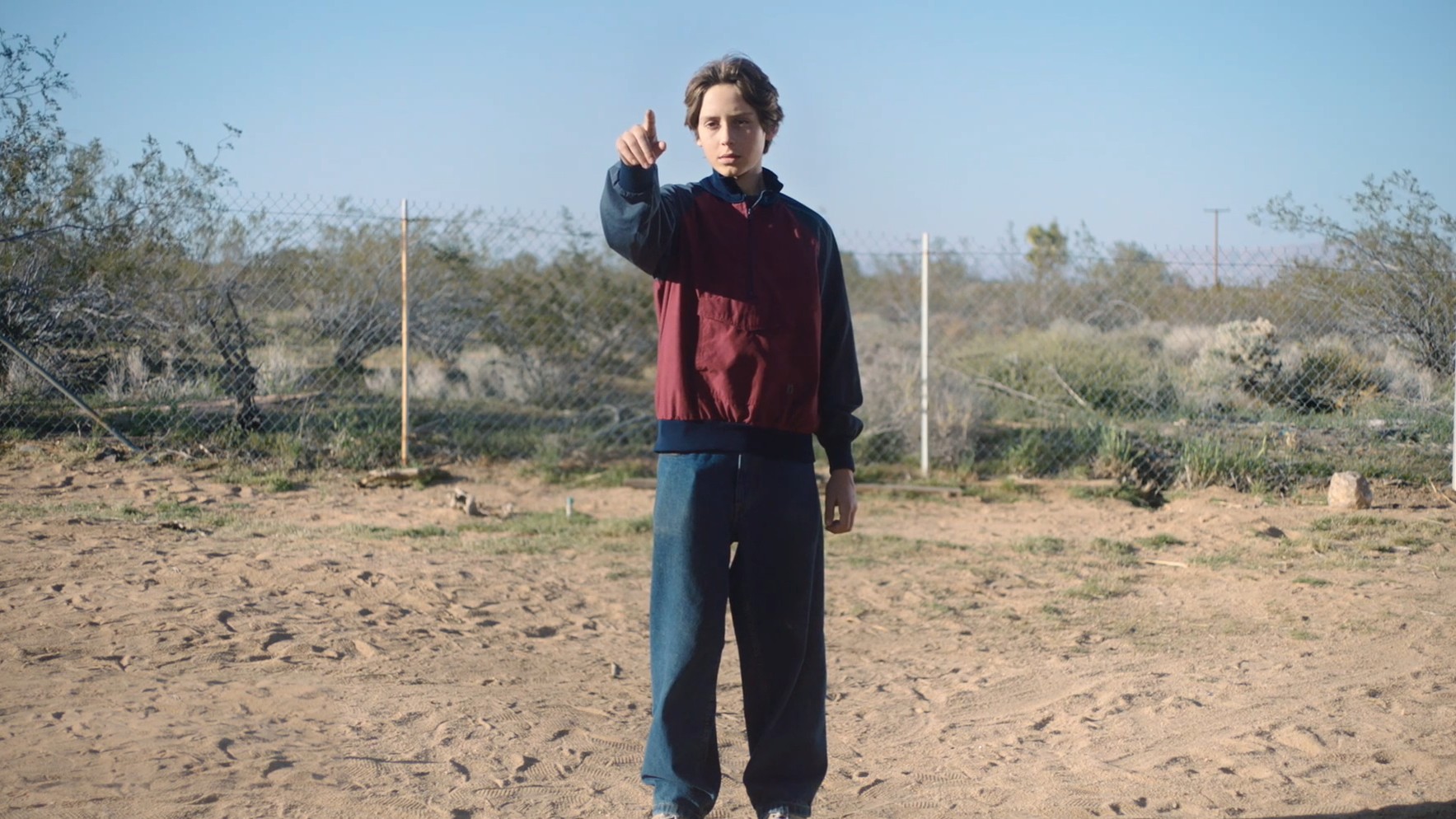 A person in a maroon and navy jacket points forward in a sandy outdoor setting.