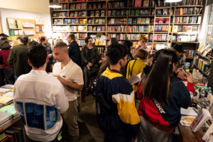 Bookshop filled with people chatting and browsing books, with shelves full of colorful titles in the background.