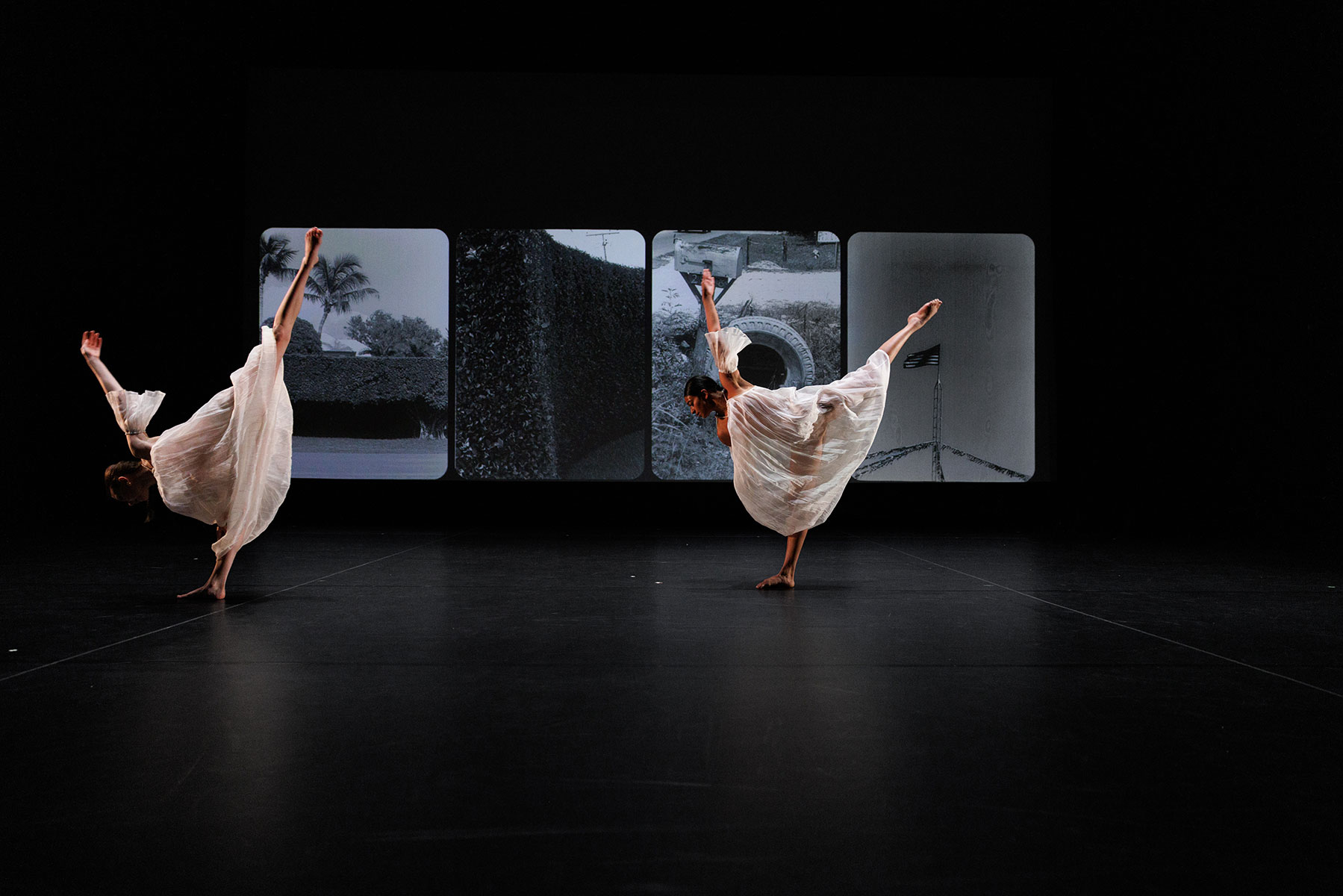Two dancers on a dark stage hold a bow like pose in front of a screen projecting old film photographs