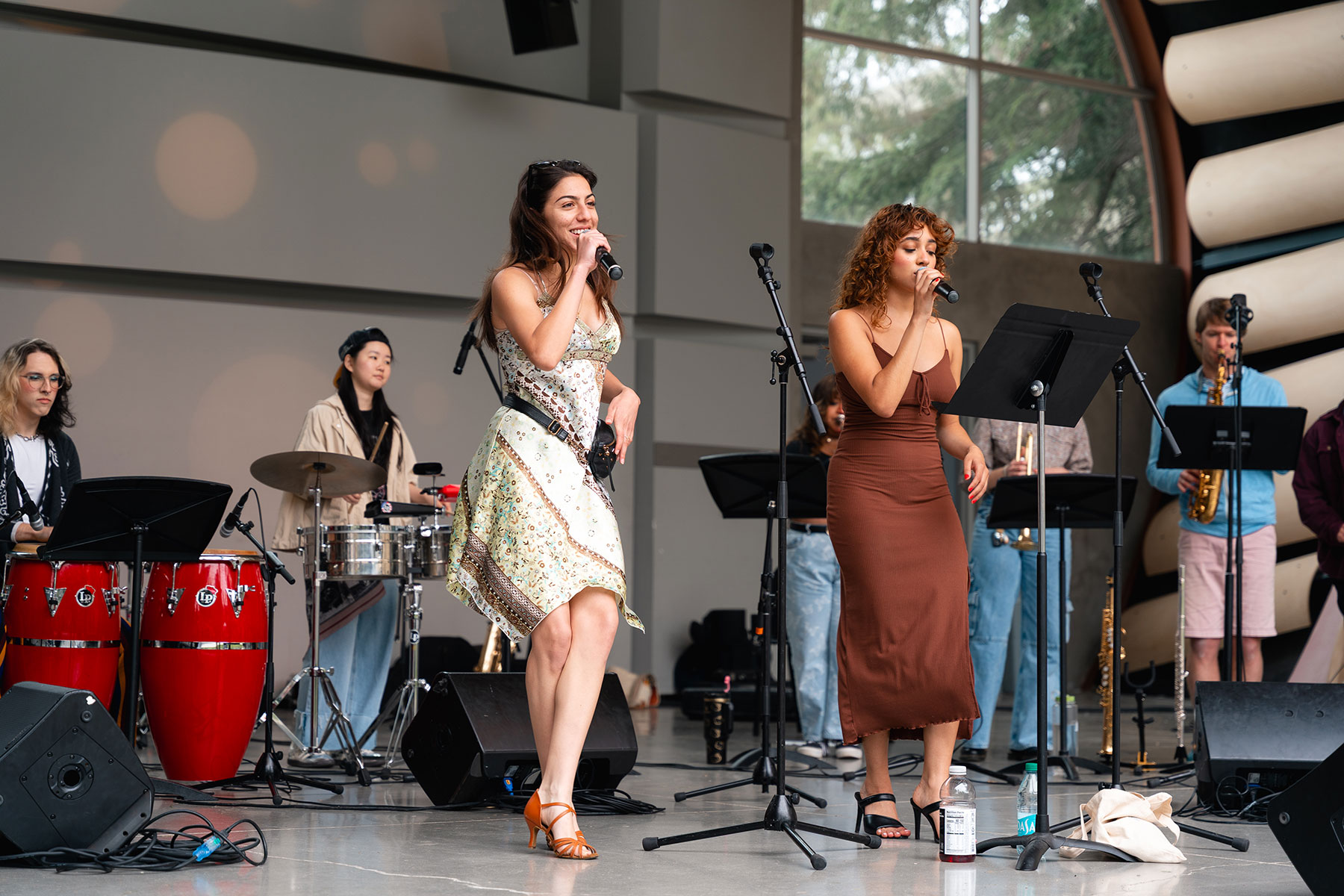 two singers hold microphones and sing while performing with a sala band including drums and brass instruments