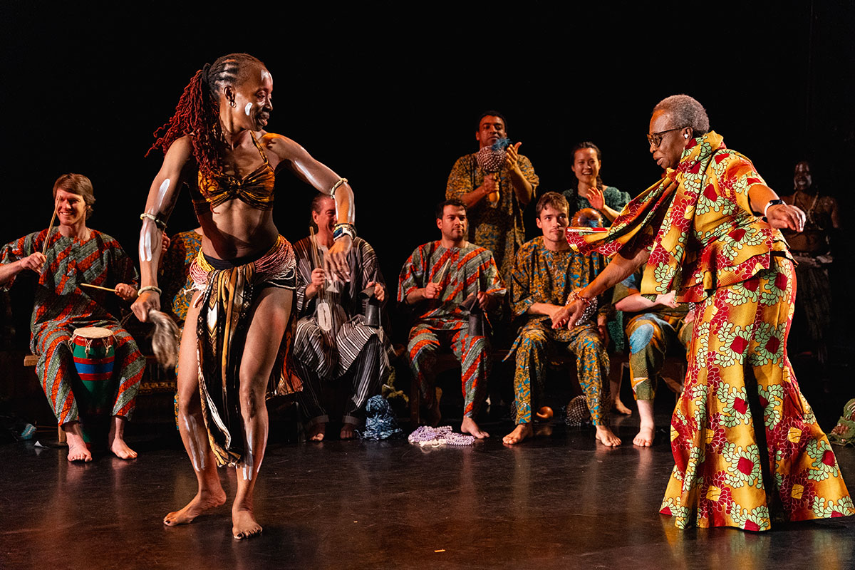 two dancers on a dark stage are dressed in traditional african clothing, behind them are a line of drummers