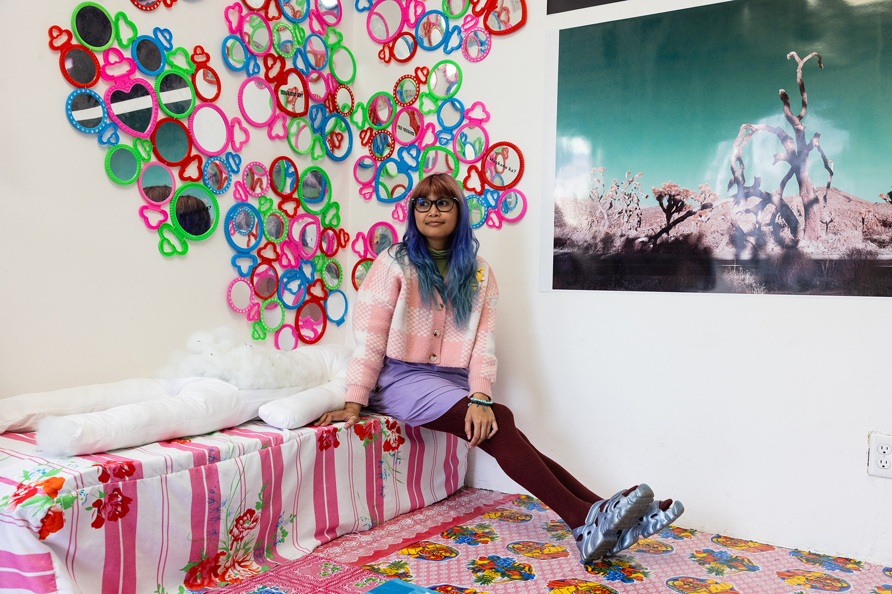 A student with blue hair sits in a brightly decorated room with a large photograph and small colorful mirrors covering the walls