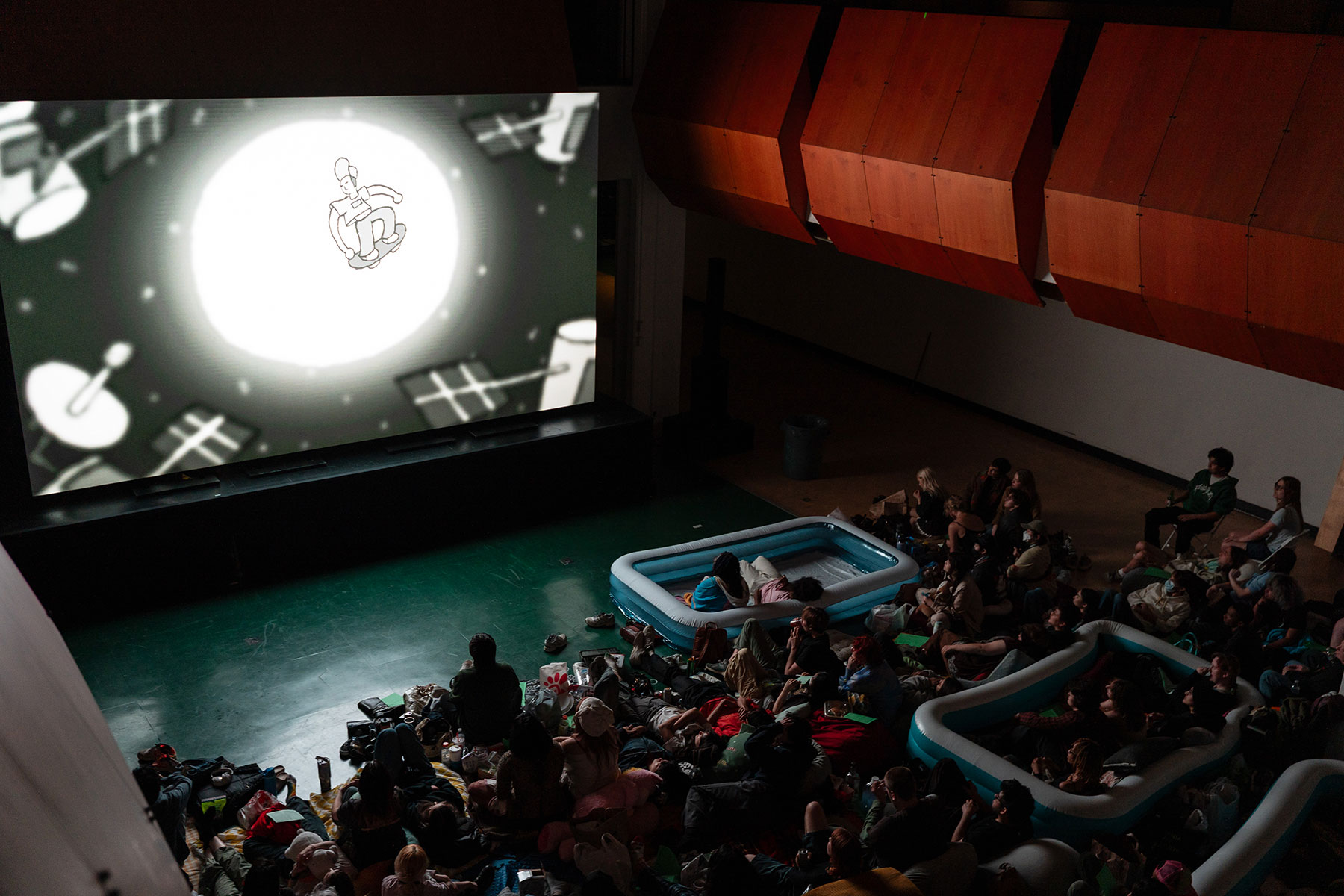 A large screen displays an animated black and white image while numerous students huddle together on the floor to watch the film