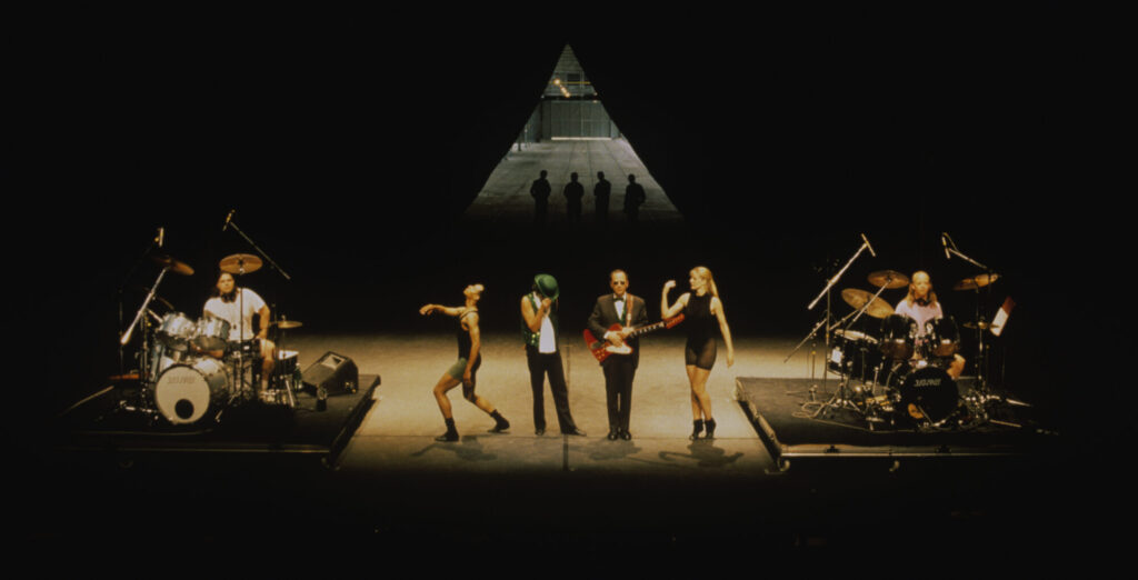 Four performers stand on stage, bookended by two drummers, with a triangle and their shadows at the back of the stage.