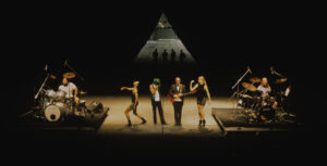 Four performers stand on stage, bookended by two drummers, with a triangle and their shadows at the back of the stage.