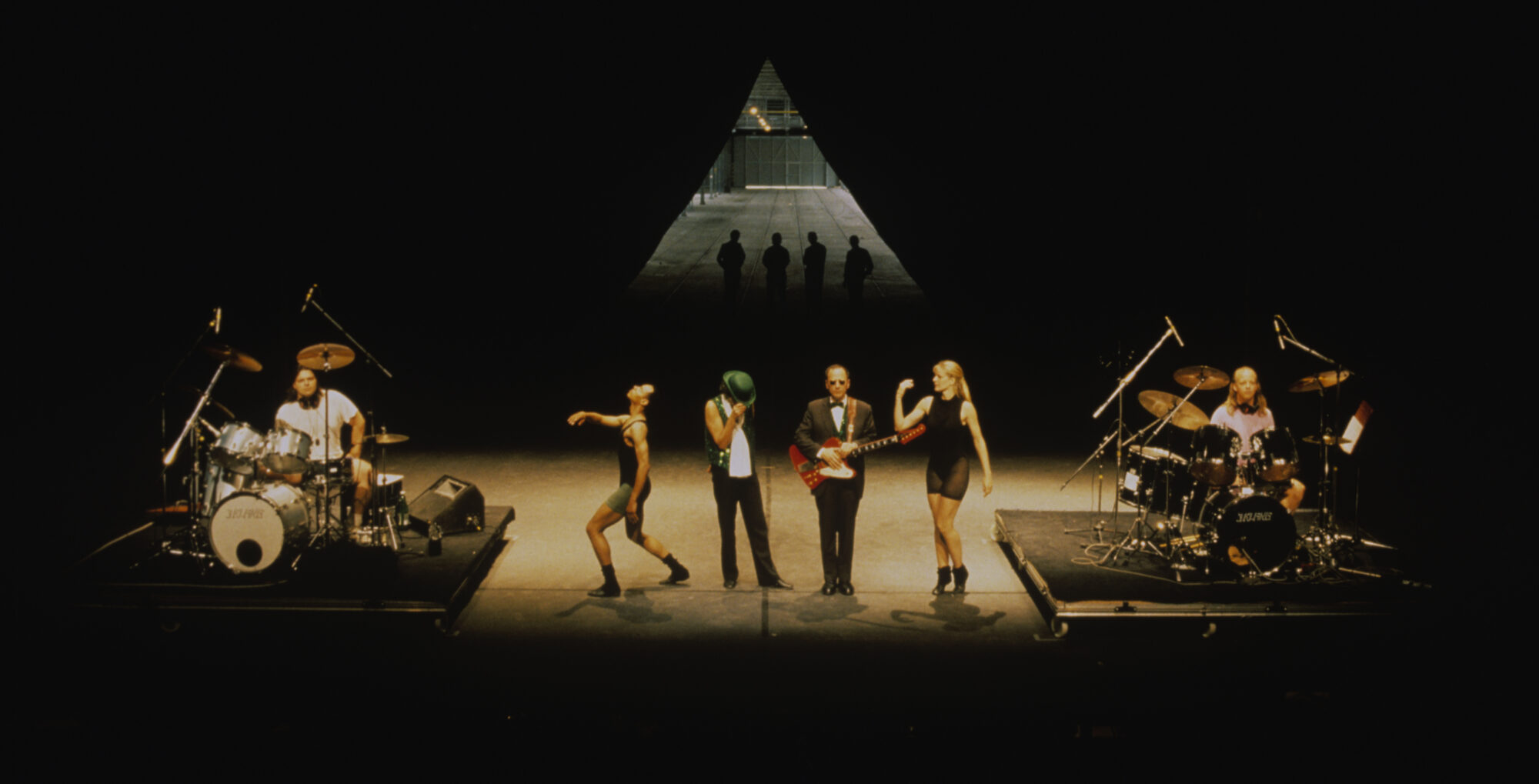 Four performers stand on stage, bookended by two drummers, with a triangle and their shadows at the back of the stage.