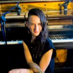 A woman in black sits at a piano, turned around and facing the camera