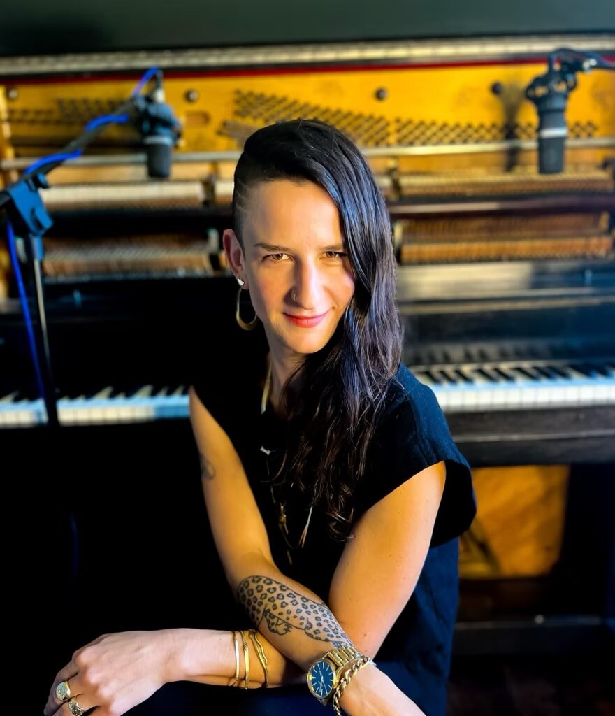 A woman in black sits at a piano, turned around and facing the camera