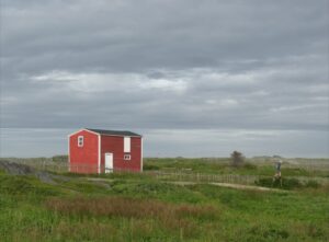 A house sits alone on a grassy plain with a person on stilts walking on a dirt road.