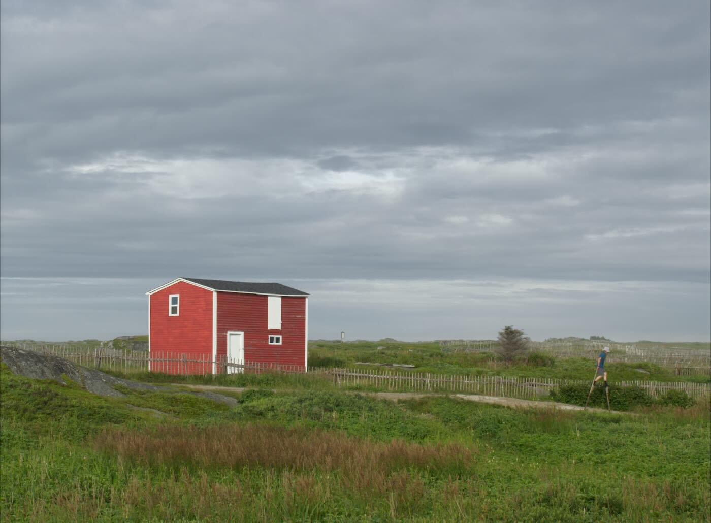 A house sits alone on a grassy plain with a person on stilts walking on a dirt road.