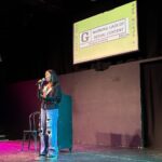A woman at a mic stands on stage underneath a screen that has a G rating.