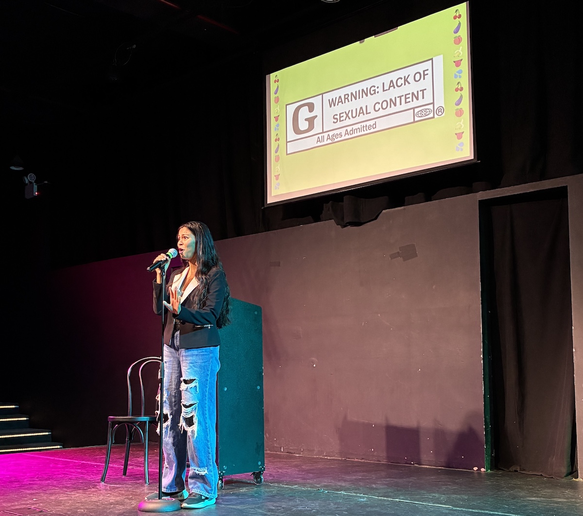 A woman at a mic stands on stage underneath a screen that has a G rating.